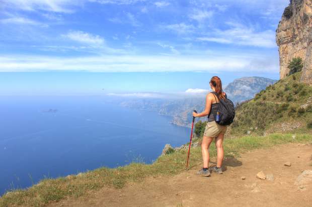 A hiker stands with a walking stick, looking over a coastal cliff. The scene features a vast ocean view under a partly cloudy sky, with rugged cliffs surrounding.