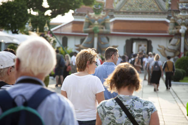 People walk towards an ornate building with a giant statue in front, under sunny skies. The scene suggests a busy, tourist environment with diverse visitors exploring the area.