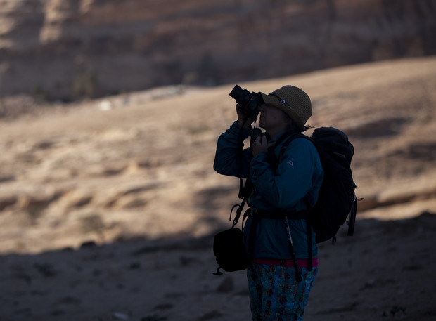 A person is photographing with a camera, wearing a backpack and hat, silhouetted against a rocky desert landscape, creating a shadowed foreground.