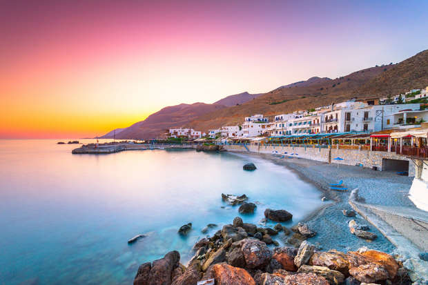 Sunset illuminates a coastal village where white buildings line a rocky shoreline. The calm sea reflects vibrant pink and orange hues, set against mountainous terrain in the background.