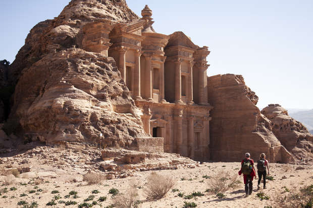 An ancient rock-carved structure stands prominently amid arid desert landscape, while two hikers approach with backpacks, emphasizing the site's grandeur and historical significance.