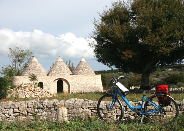 A blue bicycle rests beside a stone wall, near conical-roofed stone huts in a grassy rural landscape under a cloudy sky, with a large tree providing shade nearby.