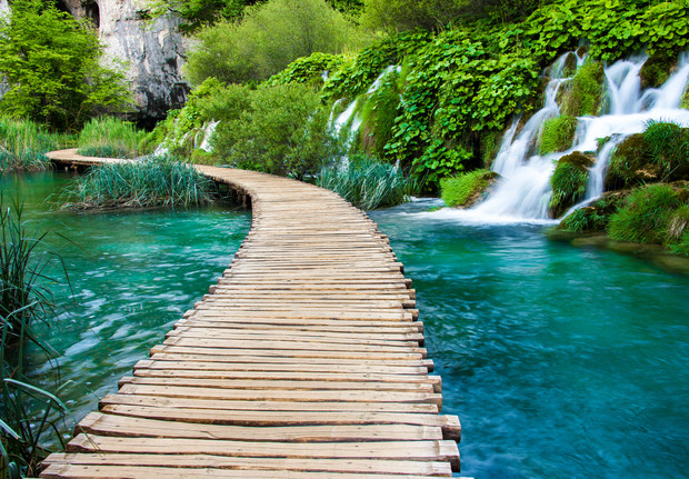 A wooden boardwalk curves over a clear turquoise river, bordered by green vegetation and small cascading waterfalls, set in a lush, natural landscape.