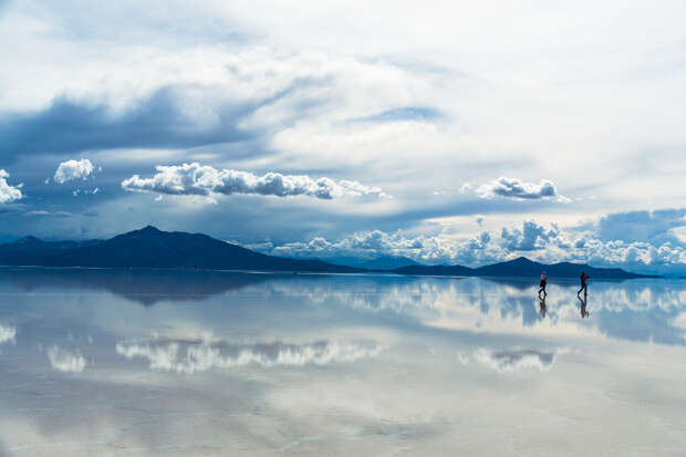 Two people walk across a vast, reflective salt flat, mirroring dramatic clouds and distant mountains under a wide, expansive sky.