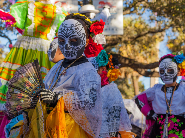 People in elaborate Day of the Dead costumes wear skull makeup and vibrant dresses while holding fans, standing outdoors during a colorful cultural celebration with trees and blue sky in the background.