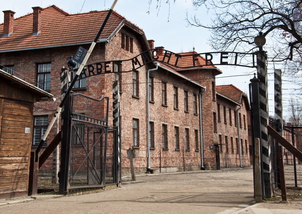 A wrought-iron gate with the phrase "ARBEIT MACHT FREI" stands open. It's part of a brick building complex, with leafless trees nearby and a clear, bright sky.