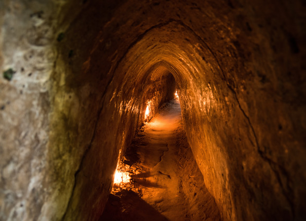 A narrow, dimly lit tunnel with earthen walls curves gently. Soft, warm lights illuminate the path, casting shadows around the enclosed, ancient underground passageway.