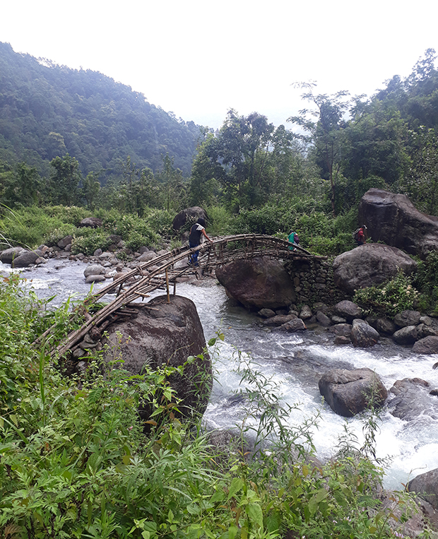 A wooden bridge spans a flowing river with people crossing it. Dense green foliage and large rocks surround the river in a hilly, forested landscape.
