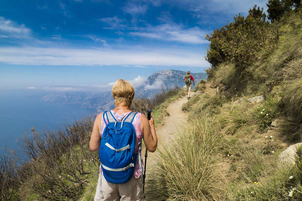 A backpacker walks along a coastal trail with trekking poles, surrounded by grassy hillsides and distant cliffs under a clear sky. Another hiker approaches in the background.