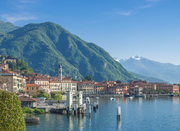 A lakeside town with pastel buildings and a clock tower sits quietly along a serene waterfront, surrounded by green mountains and distant snow-capped peaks under a clear blue sky.