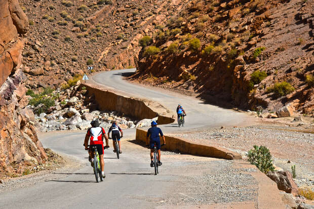 Cyclists ride through a winding mountain road surrounded by rocky terrain and sparse vegetation, under a clear, sunny sky. The path curves gently, leading deeper into the arid landscape.