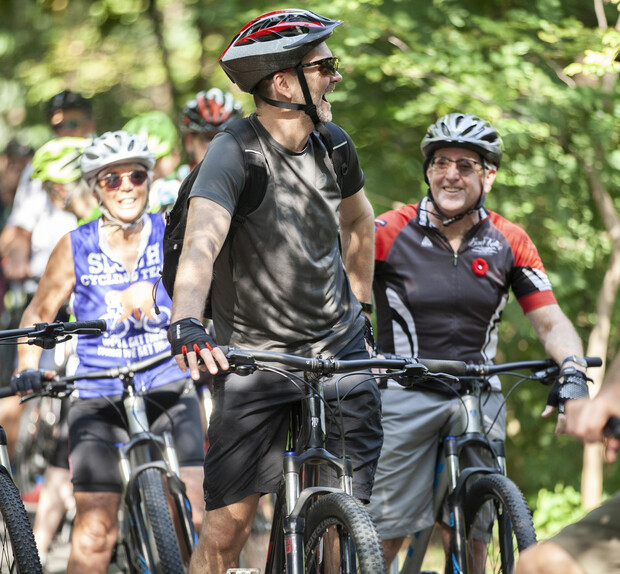 Cyclists pause and smile, wearing helmets and biking gear, surrounded by lush greenery. One shirt reads "SURVIVOR CYCLING TEAM." They enjoy a relaxed moment on a forested trail.