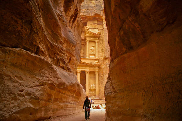 A narrow passageway features a person walking towards the intricately carved stone facade of an ancient building, surrounded by towering sandstone cliffs.