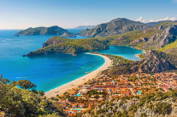 Coastal town nestled between turquoise sea and mountainous landscape; beach curves along the shore, dotted with small boats in the bay; red-roofed buildings and lush greenery create a colorful scene.
