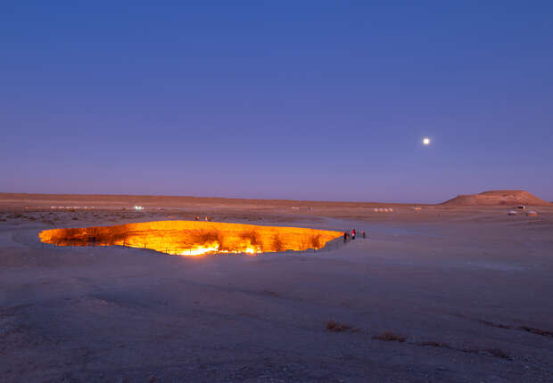 A fiery crater emits bright orange flames, illuminating the barren desert landscape under a twilight sky with a visible moon, surrounded by sparse vegetation and distant hills.