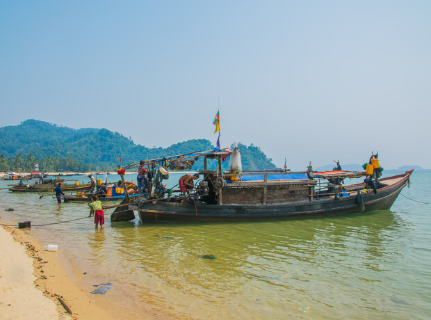 A wooden boat with colorful flags is docked on a sandy shore, surrounded by people working near a calm, shallow sea, with green hills in the distant background.