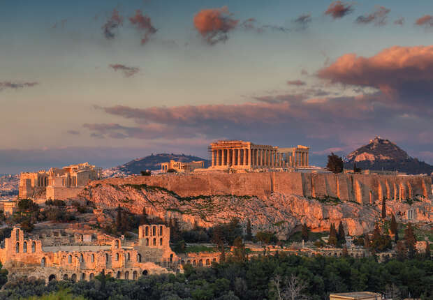 Ancient ruins, including the Parthenon, stand illuminated atop the Acropolis under a dramatic sunset sky, surrounded by hills and lush greenery, with distant city structures in the background.
