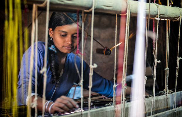 A young woman weaves fabric on a handloom, concentrating intently. Threads in various colors hang around her, while dim light provides a warm, focused atmosphere in a small workshop.