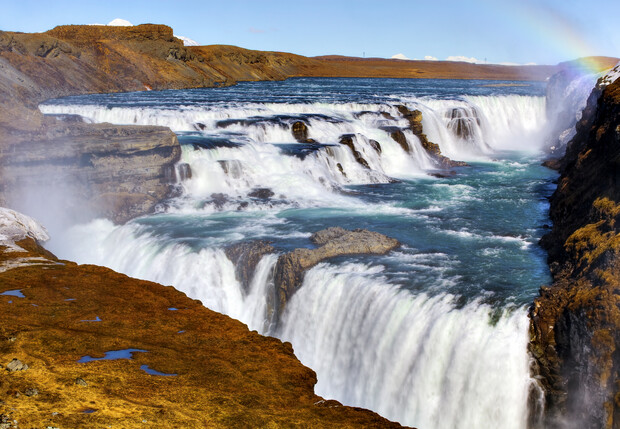 Water cascades down multiple tiers of a broad waterfall in a rocky landscape, with mist rising and a rainbow arching across a clear blue sky.