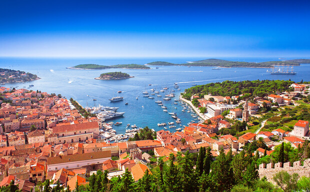 Boats are anchored in a vibrant bay, surrounded by a historic town with orange-tiled roofs and green trees. The sea is dotted with small islands under a clear blue sky.