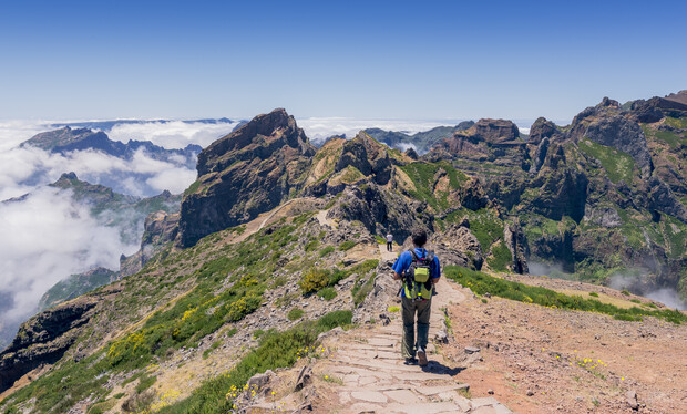 A hiker walks along a narrow mountain path surrounded by rugged, green peaks and partially covered clouds under a clear blue sky. The trail is lined with scattered vegetation.