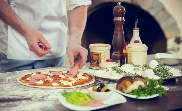A chef places ham on a pizza in a rustic kitchen, surrounded by ingredients like herbs, eggs, flour, and spices, with a visible stone oven in the background.