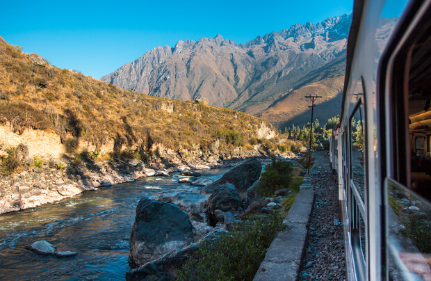 A train travels alongside a rocky river under clear blue skies, with rugged mountains in the background. The natural landscape is sunlit, showcasing the terrain's textures and colors.