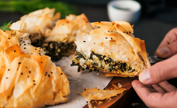 A slice of spinach-filled phyllo pastry being lifted from a wooden board, showing flaky layers and black seeds, with blurred greens and a white cup in the background.