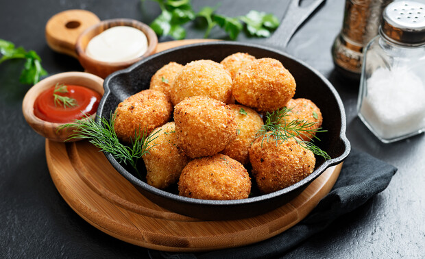 Golden fried balls stacked in a cast-iron skillet, garnished with dill, on a wooden board; accompanied by ketchup, mayonnaise, salt, and pepper, with fresh herbs in the background.