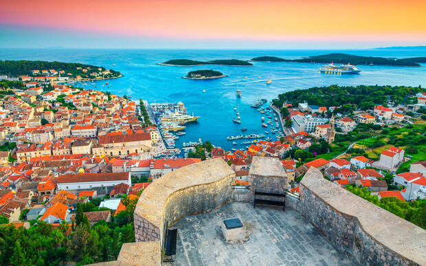 A coastal town with red rooftops overlooks a vibrant blue harbor filled with boats. The scene is set during sunset, casting warm hues over the buildings and distant islands.