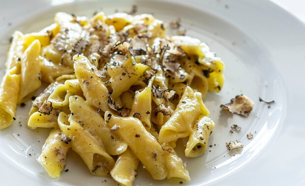 Pasta drizzled with oil and sprinkled with grated truffle sits elegantly on a white plate, presenting a gourmet dish in a simple table setting.