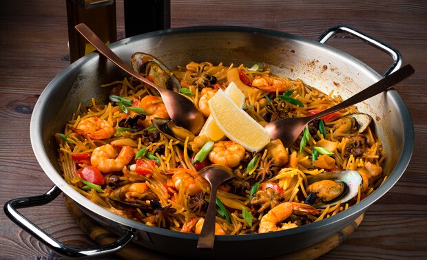 A metal pan holds a seafood pasta dish with shrimp, mussels, and vegetables, garnished with lemon wedges, on a wooden table. Three spoons rest inside, ready for serving.