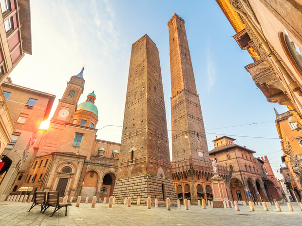 Two tall medieval towers stand prominent in an open plaza, surrounded by historic buildings with arches and a domed structure, under a clear blue sky with sunlight filtering through.