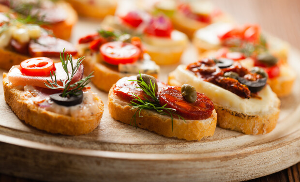 Slices of toasted bread topped with various ingredients, such as olives, tomatoes, cheese, and herbs, are arranged on a wooden platter in a casual and inviting setting.