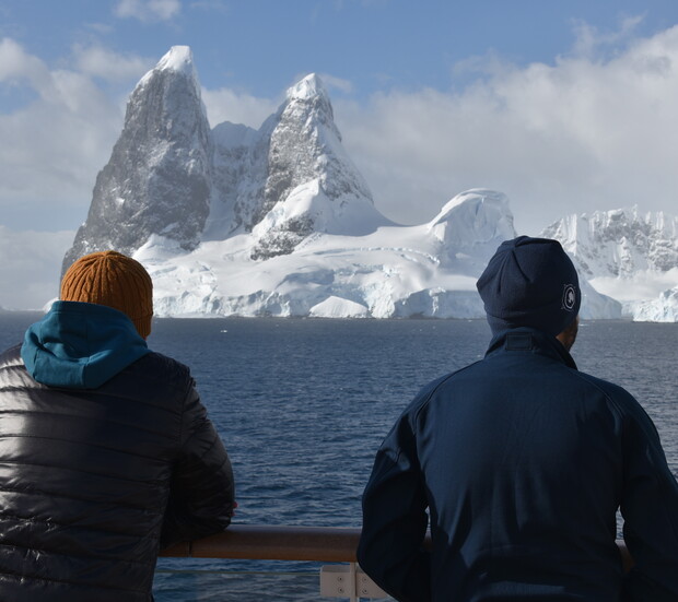 Two people in winter clothing stand on a ship's deck, looking at a massive, snow-covered mountain rising from the ocean under a partly cloudy sky.