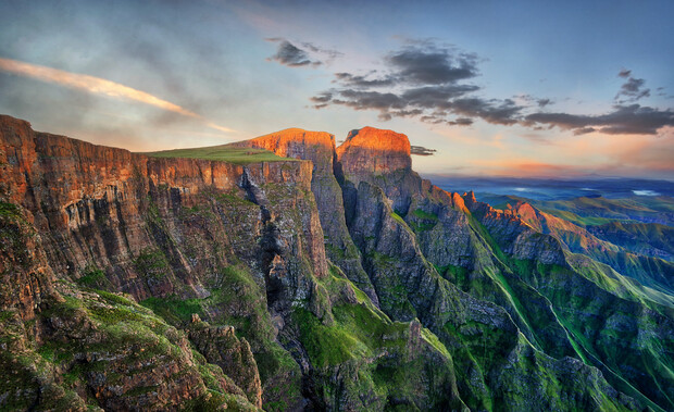 Rocky cliffs stand majestically, bathed in warm sunlight, with deep green valleys below. The sky is a mix of soft clouds and a radiant sunset, creating a tranquil, expansive landscape.