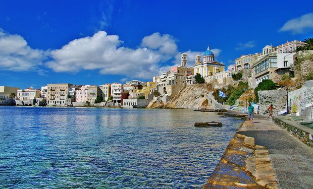 Coastal town with colorful buildings perched on a hillside. People walk along a waterfront path beside calm, clear water under a bright blue sky with scattered clouds.