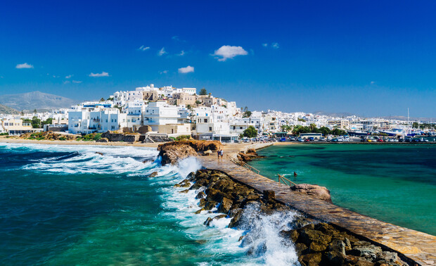 White buildings sit atop a rocky shore as waves crash against a stone walkway leading to a coastal town under a clear blue sky.