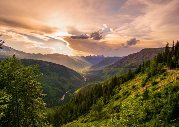 A scenic mountain valley glows under a dramatic sunset, with lush green slopes and a meandering river below, surrounded by expansive forested hills and distant peaks.