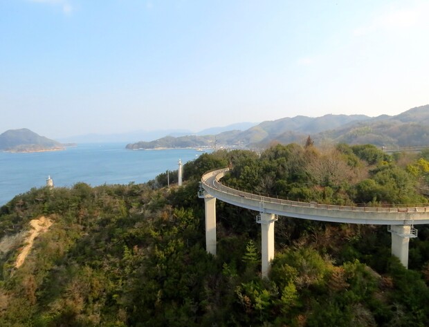 A curved road on elevated pillars winds above dense green forest, with a serene sea and distant hilly islands visible under a clear blue sky.