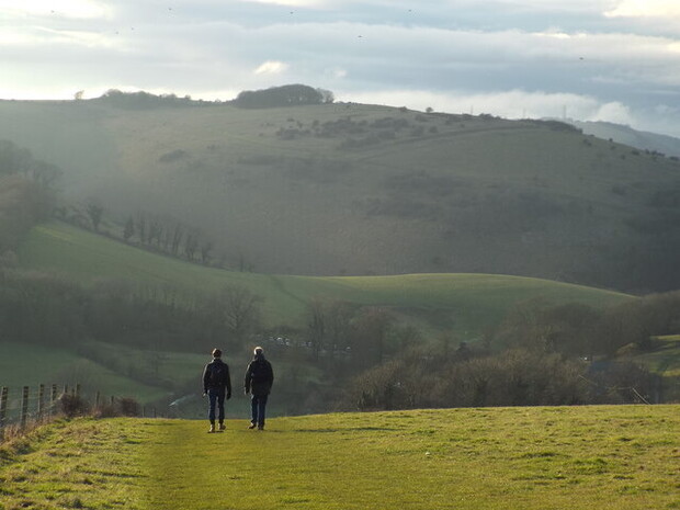 Two people walk on a grassy path surrounded by rolling hills and scattered trees under a cloudy sky, creating a peaceful rural landscape.