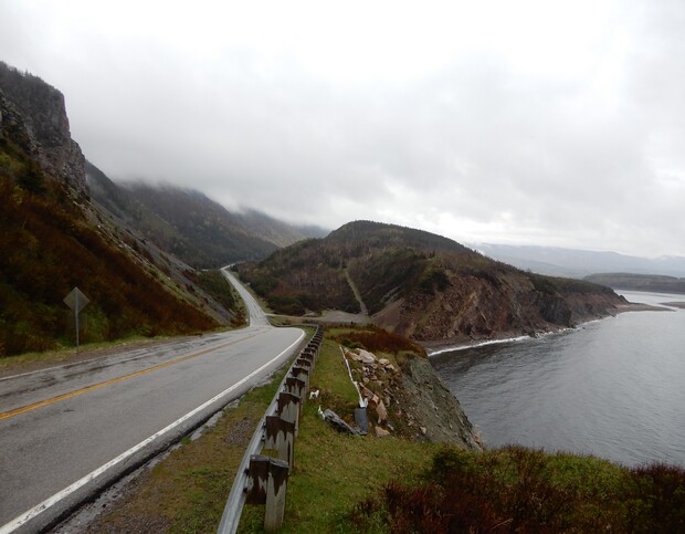 A winding road curves along a misty, coastal cliffside. The landscape appears rugged, with overcast skies and a calm body of water below, bordered by rocky terrain and sparse vegetation.