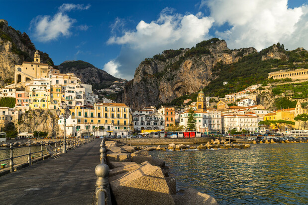 Buildings are clustered on a hillside, overlooking a waterfront path bordered by a stone railing. The scene features rocky cliffs and a blue sky with clouds in the background.