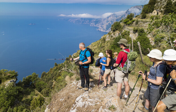 Hikers pause on a narrow, rocky trail overlooking a vast ocean with distant islands, surrounded by lush greenery and rugged cliffs under a clear blue sky.