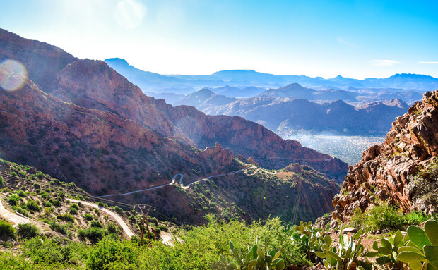Sunlit red rocky mountains stretch into the distance, with winding roads cutting through greenery in the foreground and a sprawling valley surrounded by hazy peaks under a clear blue sky.