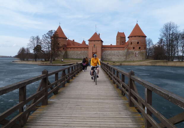 Cyclist rides on a wooden bridge towards a medieval-style castle with red-tiled turrets, surrounded by a partially frozen lake and bare trees under a clear, blue sky.