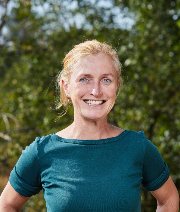 A person stands smiling with hands on hips, wearing a teal shirt. The background features lush green foliage, suggesting an outdoor, natural setting.