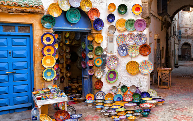 Colorful ceramic plates hang and are displayed outside a shop. The blue door contrasts with vibrant patterns. Plates cover the walls and ground, creating a lively market scene in an alleyway.