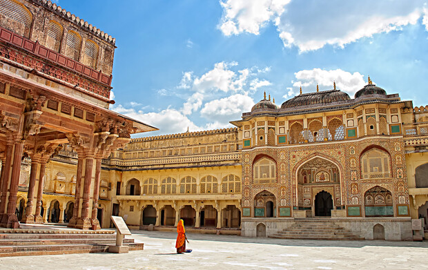 A person in vibrant attire walks across a sunlit courtyard surrounded by ornate, historical architecture with intricate carvings, under a bright blue sky filled with fluffy white clouds.