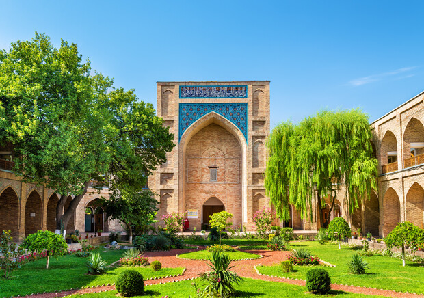 Large arched entrance stands majestically, decorated with intricate blue tile patterns. Surrounded by a serene garden with manicured paths, bordered by trees and historic brick architecture under a clear blue sky.